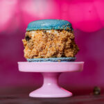 Blue macaron atop a crumb-covered cake bite on a pink pedestal, with a pink blurred background.