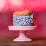 Close-up of an orange macaroon with a blue sugar-coated filling on a pink cake stand against a pink blurred backdrop