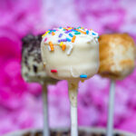 Close-up of cake pops on sticks with white chocolate coating and colorful rainbow sprinkles, against a pink blurred background.