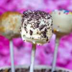 Close-up of a white chocolate-covered cake pop coated with crushed cookies, on a stick, against a pink blurred background.