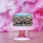 Blue macaroon with chocolate crumb topping on a pink dessert stand against a soft pink backdrop, close-up view.