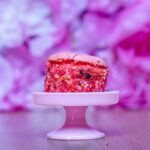 Pink macaron perched on a white pedestal against a blurred purple floral background (soft-focus).