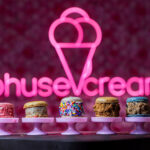 Row of pastel pink dessert stands holding six assorted donuts with colorful toppings, neon pink sign in background.