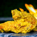 Crispy fried chicken pieces stacked on a metal surface with orange flame in the background, suggesting open-flame cooking.