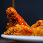 Close-up of a glossy, orange-glazed fried chicken wing lifted from a plate against a dark background.