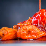 Close-up of glossy orange hot sauce pouring over crispy fried chicken wing, catching the light.