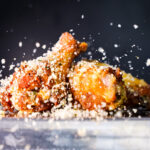 Close-up of crispy fried chicken pieces covered in seasoned breadcrumbs and salt flakes, with crumbs mid-air against a dark background.