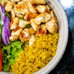 Grilled chicken pieces over yellow quinoa with greens, tomato slices, red onion, and cucumber ribbons in a bowl.