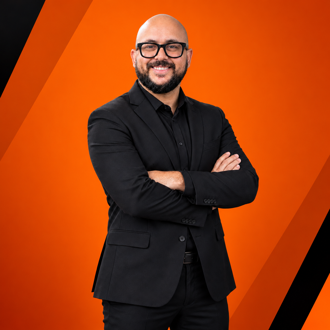 Portrait of a smiling bald man with glasses, wearing a black suit with arms crossed, against an orange geometric background.