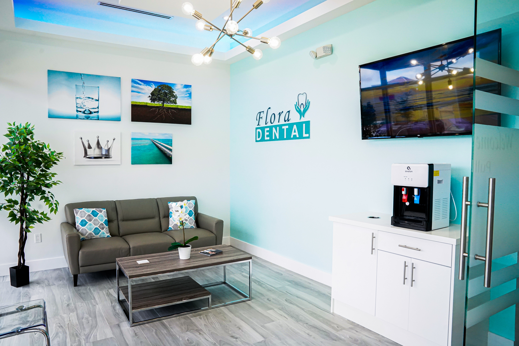 Reception area of Flora Dental with turquoise wall, logo, and a gray sofa set; wall art, plant, and modern chandelier visible.