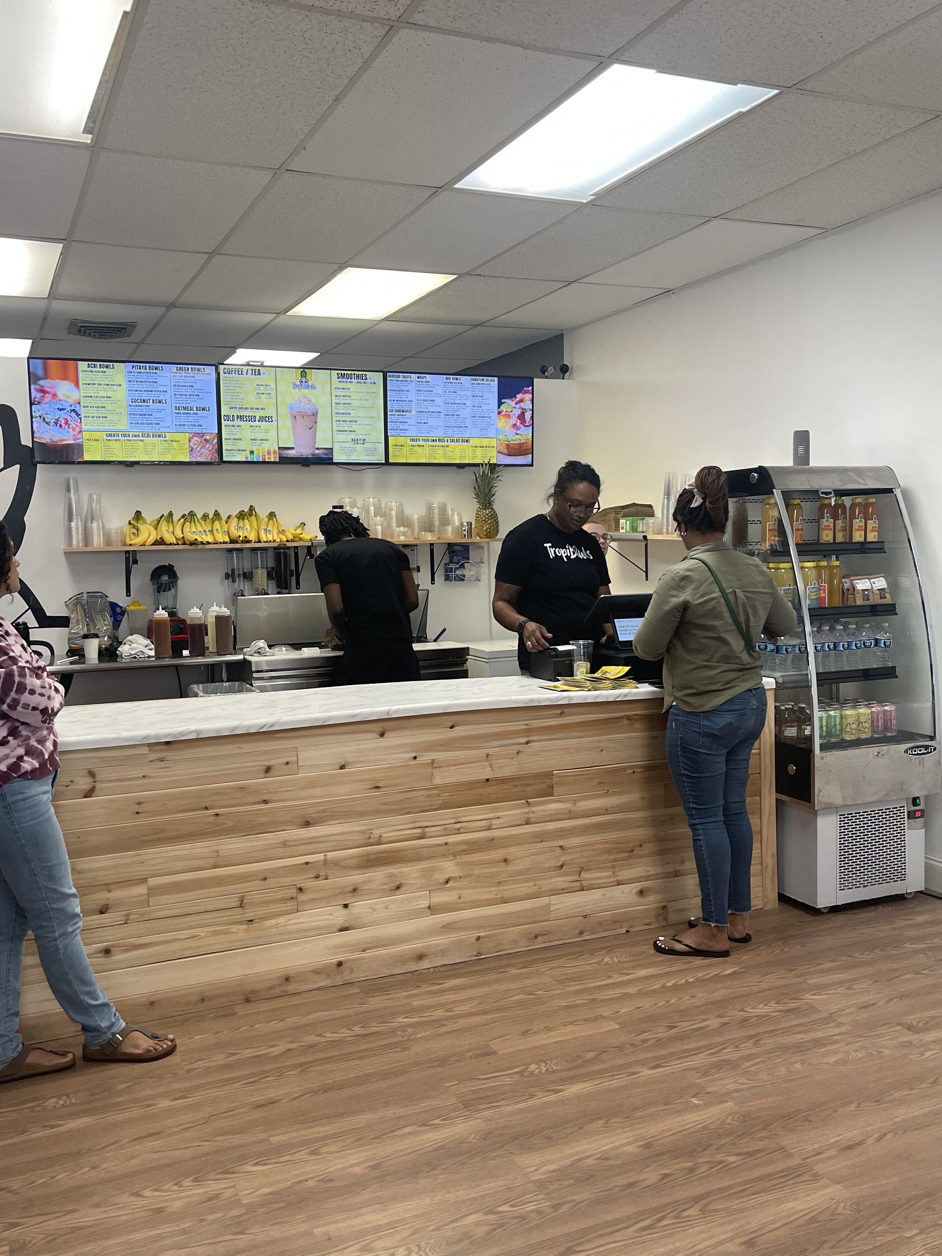 Juice bar interior with a wooden counter, menu boards overhead, bananas on a shelf, and customers at the register as staff prepare orders.