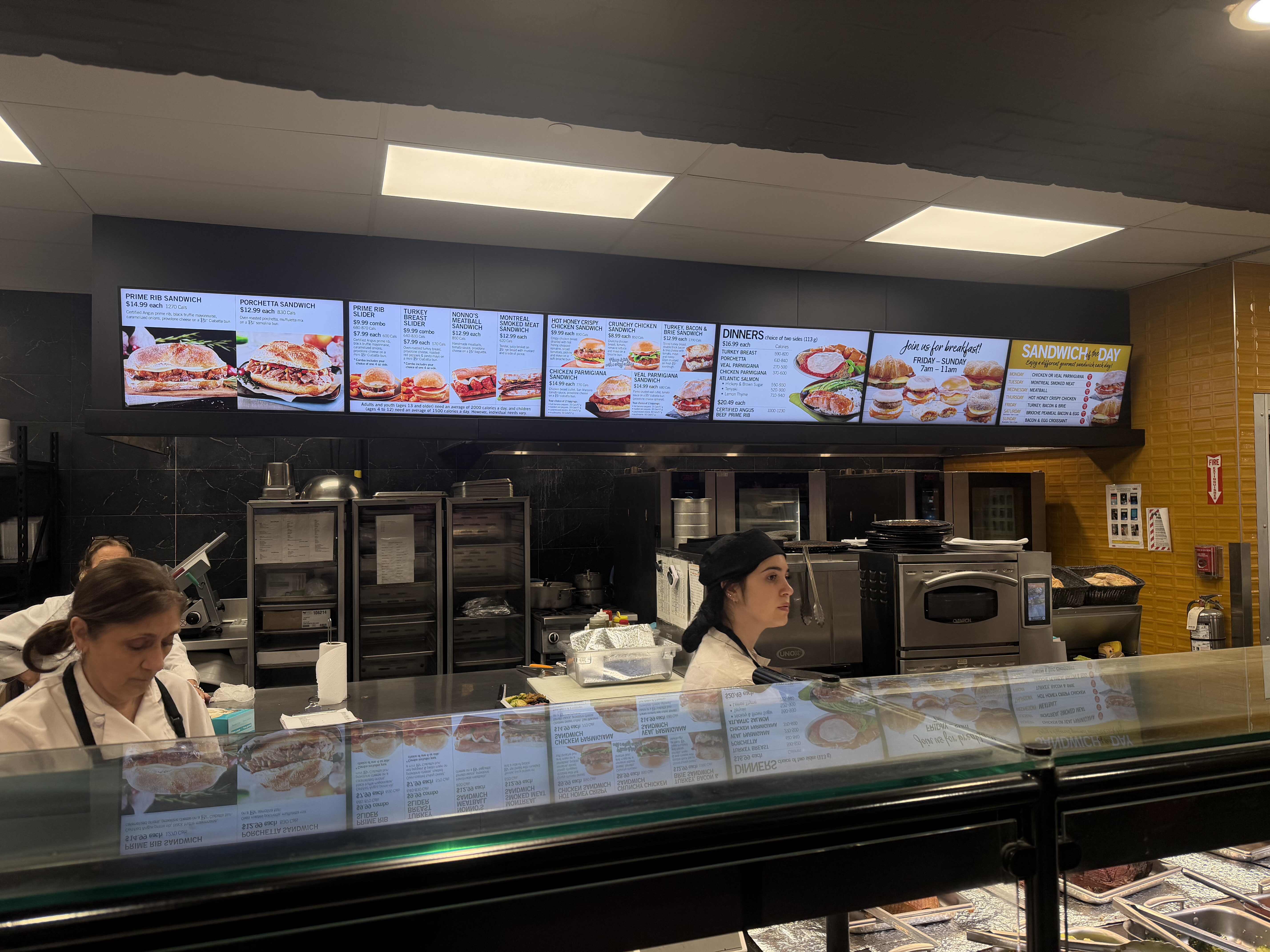 Fast-food counter with illuminated menu boards displaying sandwiches, two staff in white uniforms behind a glass display case.