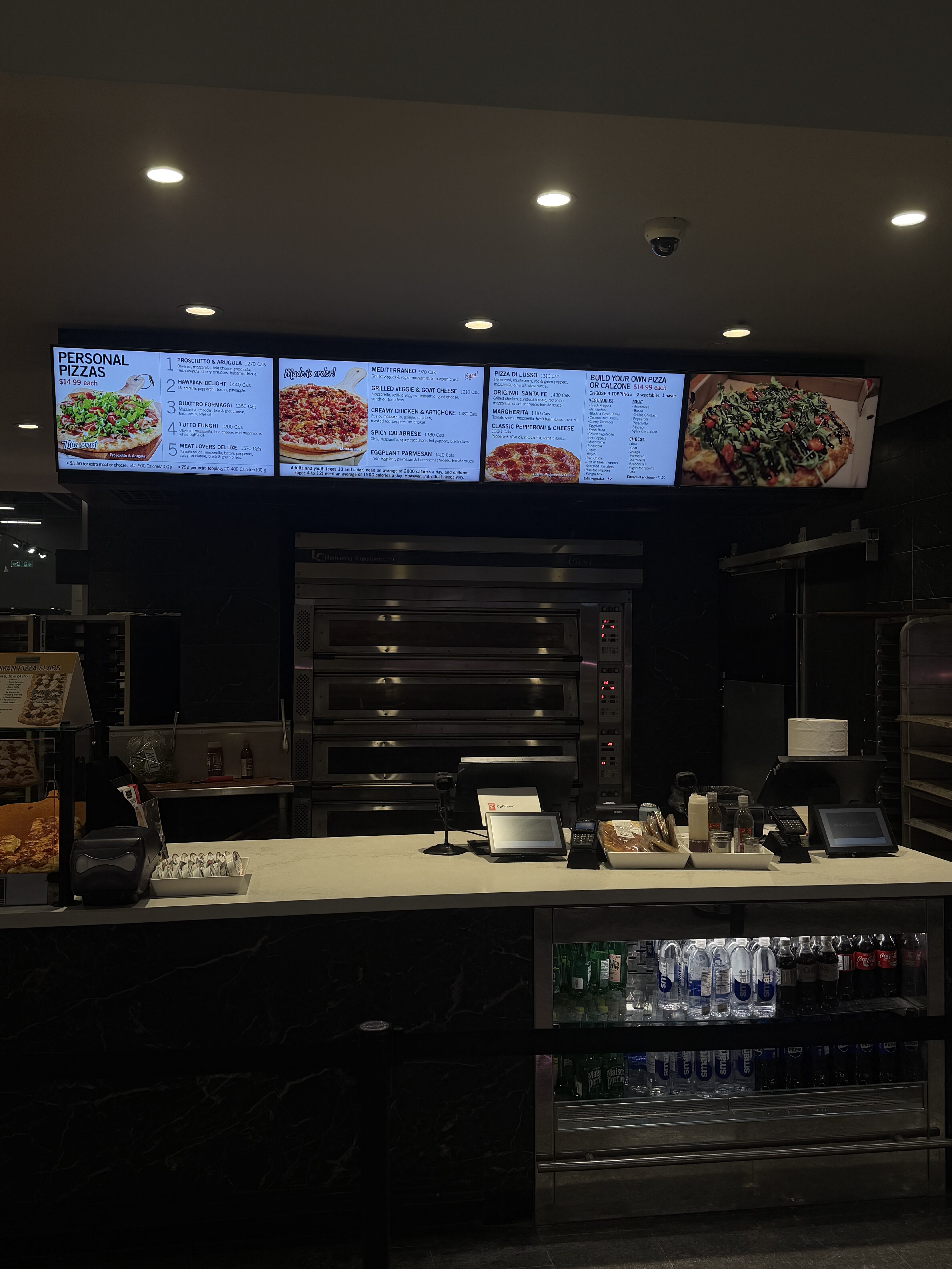 Pizza shop counter with multiple digital menu boards overhead and stainless ovens in the back; drinks cooler visible on the right side.
