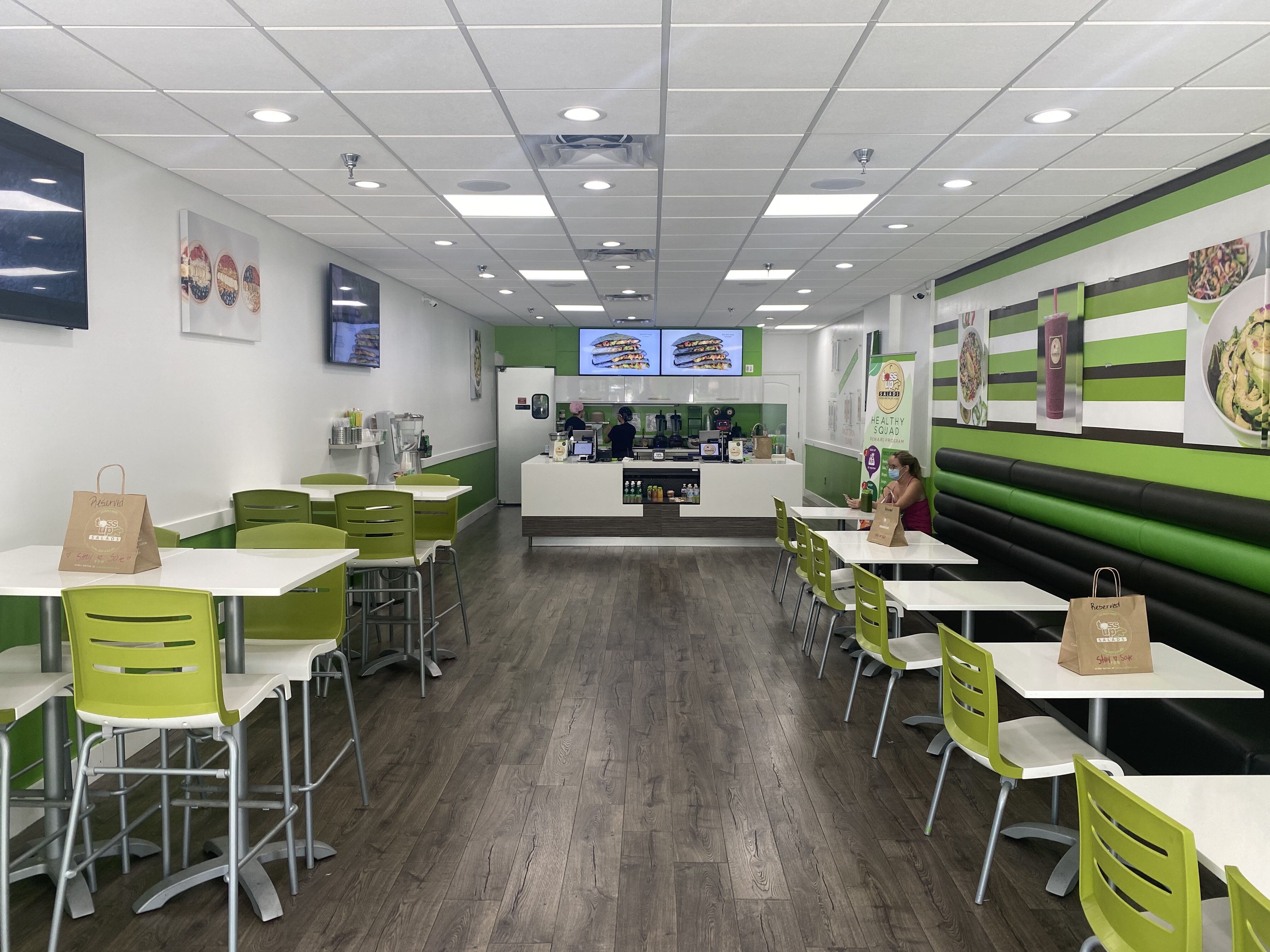 Bright fast-casual restaurant interior with white tables, lime-green chairs, and a striped green/black wall; service counter in the back with staff.