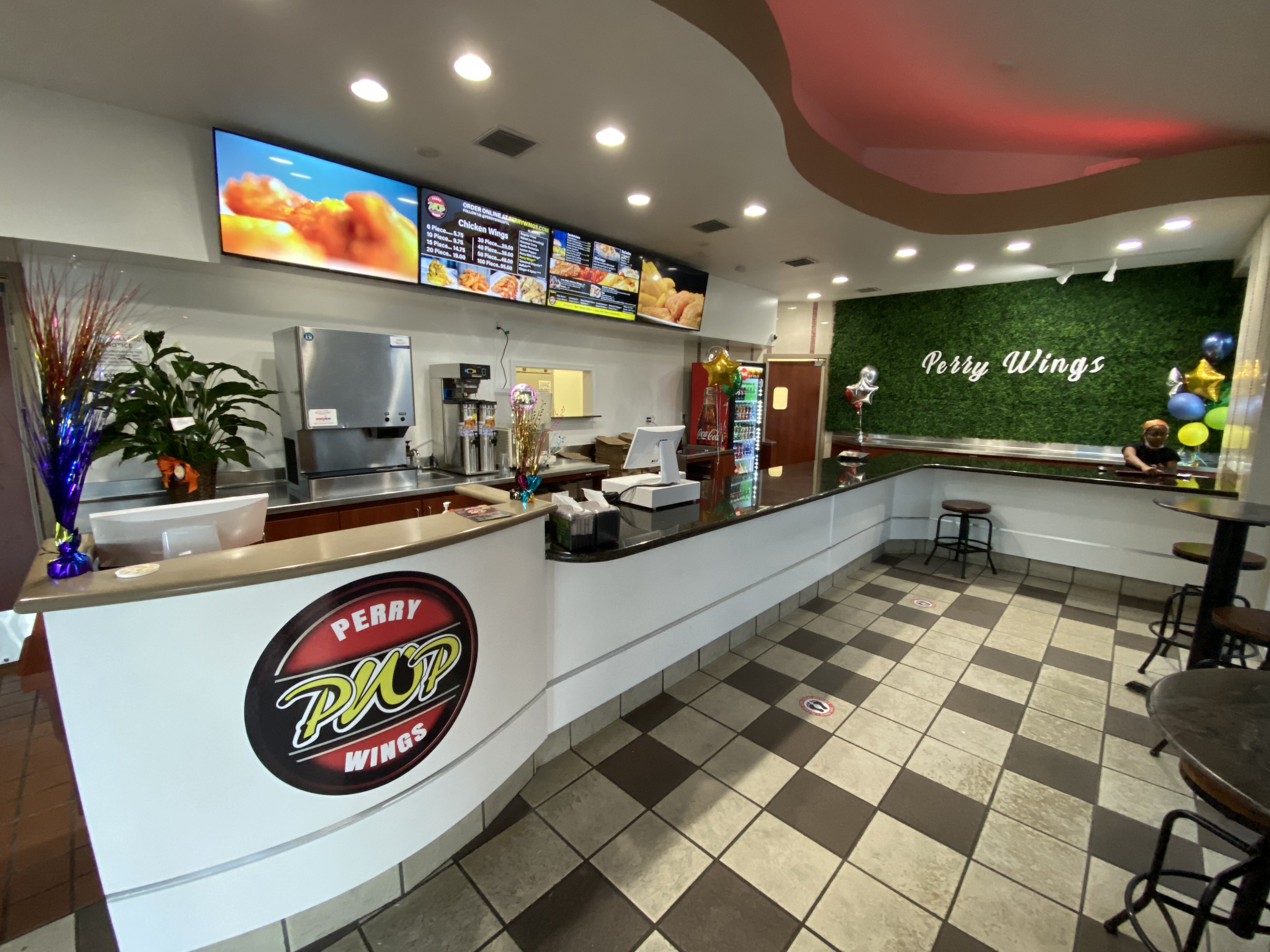 Restaurant counter with Perry Wings logo, overhead menu boards, and a soda fridge in a bright eatery setting.